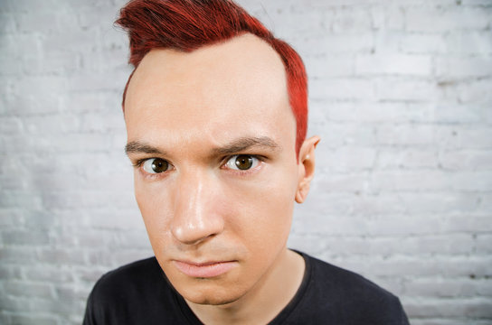 Young red-haired guy bend one's brows and looks forward on white brick wall background.