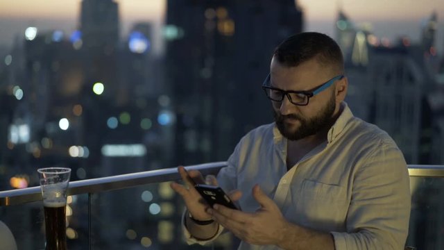 Man sitting in skybar at night and browsing internet on smartphone
