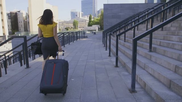 Back view of young adult asian business woman in pencil skirt walking with suitcase to airport. Business lady travelling to the airport on a summer day. Female with luggage going on business travel