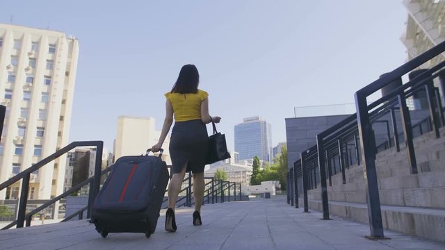Low angle back view of business woman walking on high heels with luggage. Corporate female arrived on business trip and walking with suitcase in the city business area