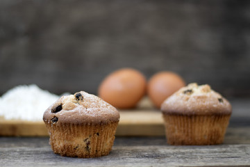 Two muffins with raisins sprinkled with powdered sugar on a wooden background and are ingridient on the back of a blurry background
