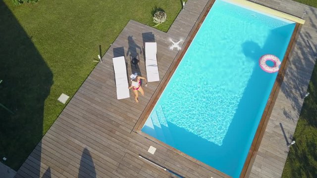 AERIAL: Woman lying by the pool gets angry at spying drone and tries to crash it. Annoying neighbor flies his drone to film woman in bikini sunbathing, who tries to bring it down by throwing things.
