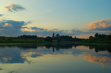Fototapeta na wymiar Church in the distance in the forest near the river against the green banks and blue sky with clouds in the evening in the summer reflected in the water