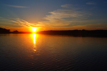 Obraz premium Beautiful amber sunset in summer over the river - orange sun with feathery clouds and blue sky reflected in the water on the background of the forest on the far shore