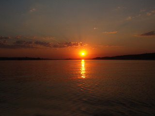 Naklejka premium A picturesque summer sunset over the water of the lake, bright orange amber sun on the horizon against the background of calm water and evening sky with a small cloud