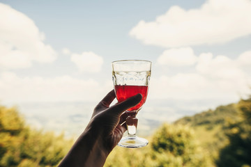 hand holding cranberry juice in glass cup on background view of mountains and sky. stylish red drink on porch. summer vacation and relax concept. travel and wanderlust. space for text