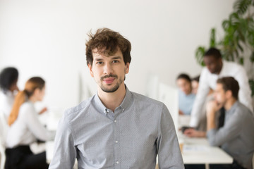 Skilled young professional looking at camera posing in office with team colleagues at background,...
