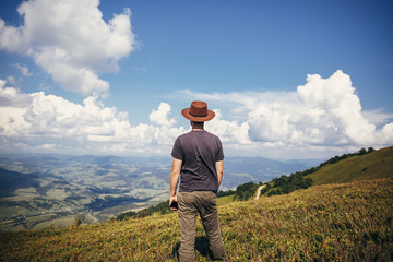 stylish traveler man in hat holding phone, walking on top of sunny mountains and sky. travel and wanderlust concept. space for text. happy hipster traveling. summer vacation