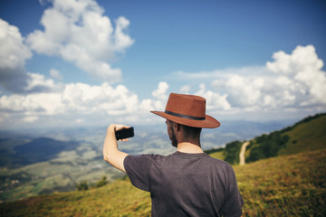 stylish traveler man in hat holding phone, walking on top of sunny mountains and sky. travel and wanderlust concept. space for text. happy hipster traveling and taking photo.