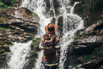 stylish hipster girl in hat with backpack looking at waterfall in forest in mountains. traveler woman exploring woods. travel and wanderlust concept. summer vacation, space for text