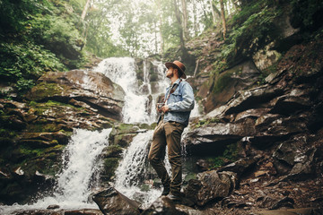 stylish hipster man in hat with photo camera, standing at waterfall in forest in mountains. traveler guy exploring woods. travel and wanderlust concept. atmospheric moment