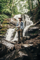 Obraz premium travel and wanderlust concept. stylish hipster man in hat with photo camera, standing at waterfall in forest in mountains. traveler guy exploring woods. space for text