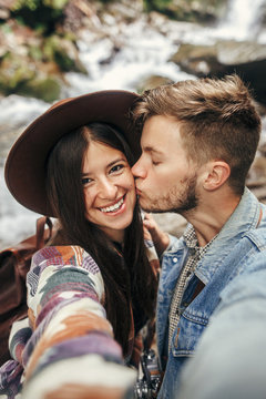 stylish hipster couple taking selfie and kissing at waterfall in forest in mountains. handsome man and happy woman in hat embracing on honeymoon. travel together. summer vacation