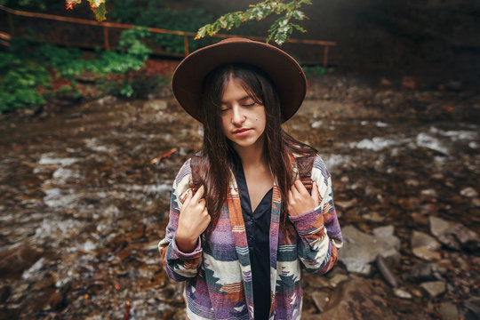 stylish hipster girl in hat with backpack relaxing in forest in mountains. traveler woman exploring summer woods. travel and wanderlust concept. atmospheric moment, space for text