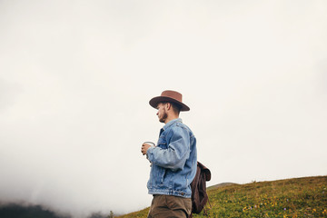 Naklejka na ściany i meble travel and wanderlust concept. stylish traveler man in hat with backpack holding cup of tea in mountains with clouds. space for text. hipster guy traveling. amazing atmospheric moment