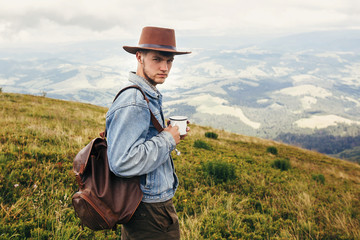 Naklejka na ściany i meble stylish traveler man in hat with backpack holding cup of tea in mountains with clouds. space for text. hipster guy traveling. amazing atmospheric moment. travel and wanderlust concept