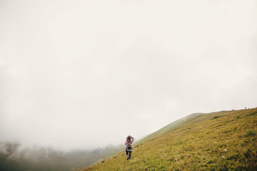 stylish traveler girl holding hat with backpack and windy hair, walking in mountains clouds. summer vacation. travel and wanderlust concept. space for text. amazing atmospheric moment