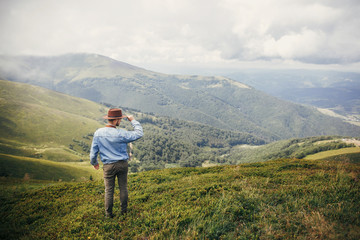 Naklejka na ściany i meble stylish traveler man in hat standing on top of sunny mountains in clouds. space for text. hipster guy traveling. amazing atmospheric moment. travel and wanderlust concept