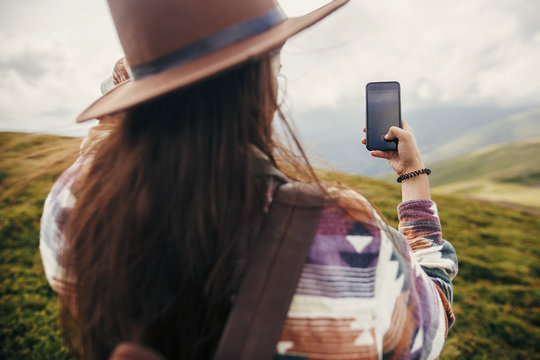 stylish traveler girl in hat with windy hair holding phone and taking picture on top of sunny mountains. travel and wanderlust concept. space for text. back view. amazing atmospheric moment