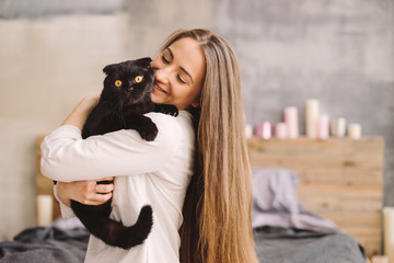 Happy young beautiful caucasian woman smiling and holding a black cat. Playing with pet at home. Love, coziness, leisure, animal protection concept. Scottish fold breed.