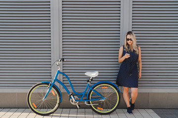 Lifestyle and technology. Young pretty blonde woman using her smartphone standing with her vintage bike on urban wall background. Good day for a ride. Copy space.
