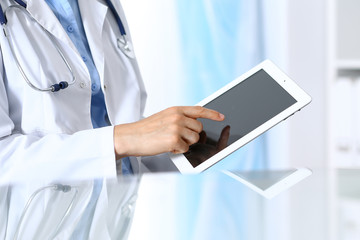 Woman doctor using tablet computer while sitting. Reflecting glass table is a working place of physician. Healthcare, insurance and medicine concept