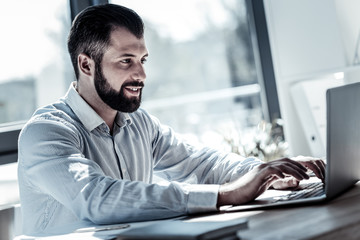 Obraz premium Im busy. Concentrated pleasant beardful man sitting in the office looking at the screen and using his laptop.