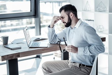 Obraz premium I must do this. Exhausted stylish responsible employee sitting in the office by the table holding the glasses in the left hand and feeling bad himself.
