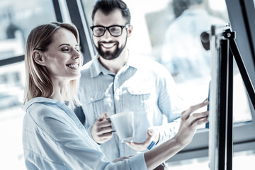New project. Satisfied pleasant occupied woman standing in the office with a man near the board focusing on her task and smiling.