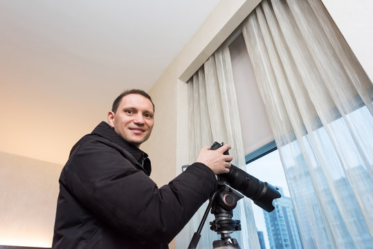 Young happy smiling man photographer with tripod taking pictures inside room of hotel, apartment building through window in New York City, NYC urban