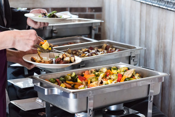 Hot buffet tray, fresh grilled vegetables with closeup of woman using tongs to serve food to plate in banquet, wedding, or restaurant inside