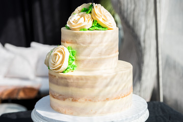 Macro closeup of three tiered vanilla golden light white yellow cream sponge wedding cake with rose flowers decoration on bright stand, green leaves