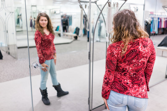 Young woman trying on clothing in boutique store looking at herself in mirror with red shirt, jeans, and boots