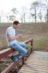 Lake Fairfax Park in winter in Reston, Virginia with young man sitting at sunset on bridge railing looking at smartphone cell phone mobile in Northern VA, texting happy smiling