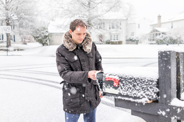 Young man checking mail in neighborhood road with snow covered ground during blizzard white storm, snowflakes falling in Virginia suburbs, single family homes in mailbox box