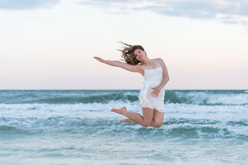 Young happy smiling woman in white dress on beach pink sunset in Florida panhandle with wind, ocean waves, jumping mid-air with water drops