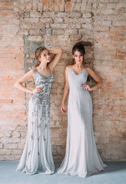 Two young girls in gray, long, shining, evening dresses with rhinestones and stones. The image of schoolgirls at the prom. Festive make-up nudes and smokies eyes. High, collected hairstyles.