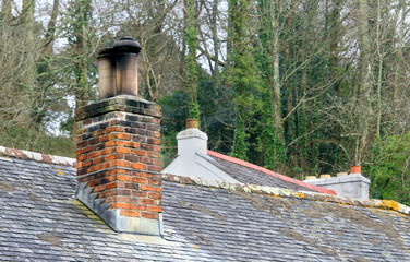 Old red brick chimney stack on a slate roofed building, with another old roof behind it and trees...