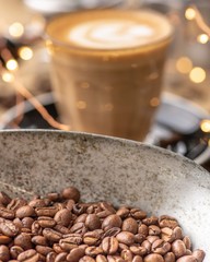 Some coffee beans in a metal scoop with a cup of coffee in the background