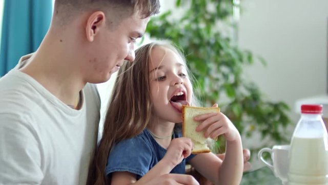 Medium shot of cute little girl licking peanut butter of toast as father laughing and talking