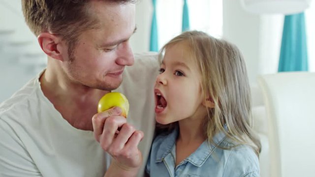 Medium shot of happy father playing with cute little daughter: they laughing and biting green apple