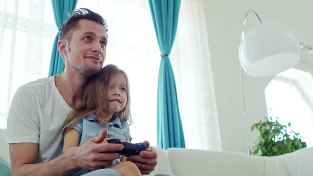 Low angle shot of cute little girl sitting on laps of happy father and playing on video game console