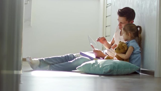 Handheld side view of happy father and cute little daughter with plush toy sitting on floor and playing on tablet