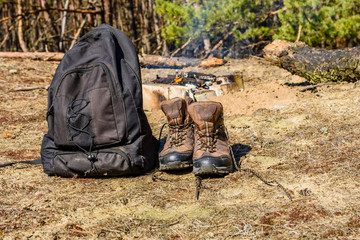 Backpack and touristic boots on a ground in a coniferous forest. Bonfire on a background