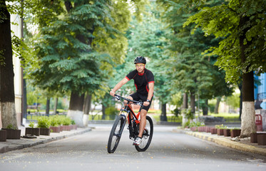 Male professional cyclist wearing cycling sportswear and protective gear riding bicycle along paved city street among green trees. Sportsman training outdoor, improving skills, exercising on fresh air