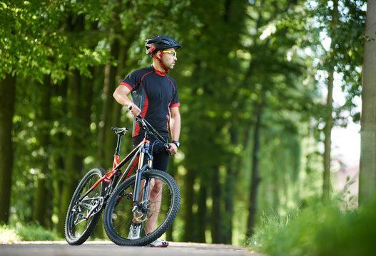 Man professional cyclist in cycling clothing and helmet looking away in distance, taking break from riding on bicycle in green park. Sportsman training outdoors, resting after race, admiring nature