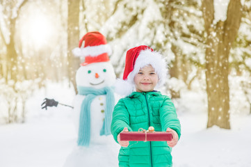 portrait of smiling boy in red christmas hat holds gift box with snowman on background