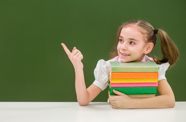 Girl with books points on empty green chalkboard and looking away
