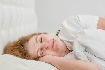 Sad sick senior woman with thermometer in mouth sleeping on the bed