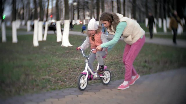 A laughing, smiling mother pushes her daughter forward on a warm spring day, when she teaches her to ride a bike along the city's sidewalk near a green park.